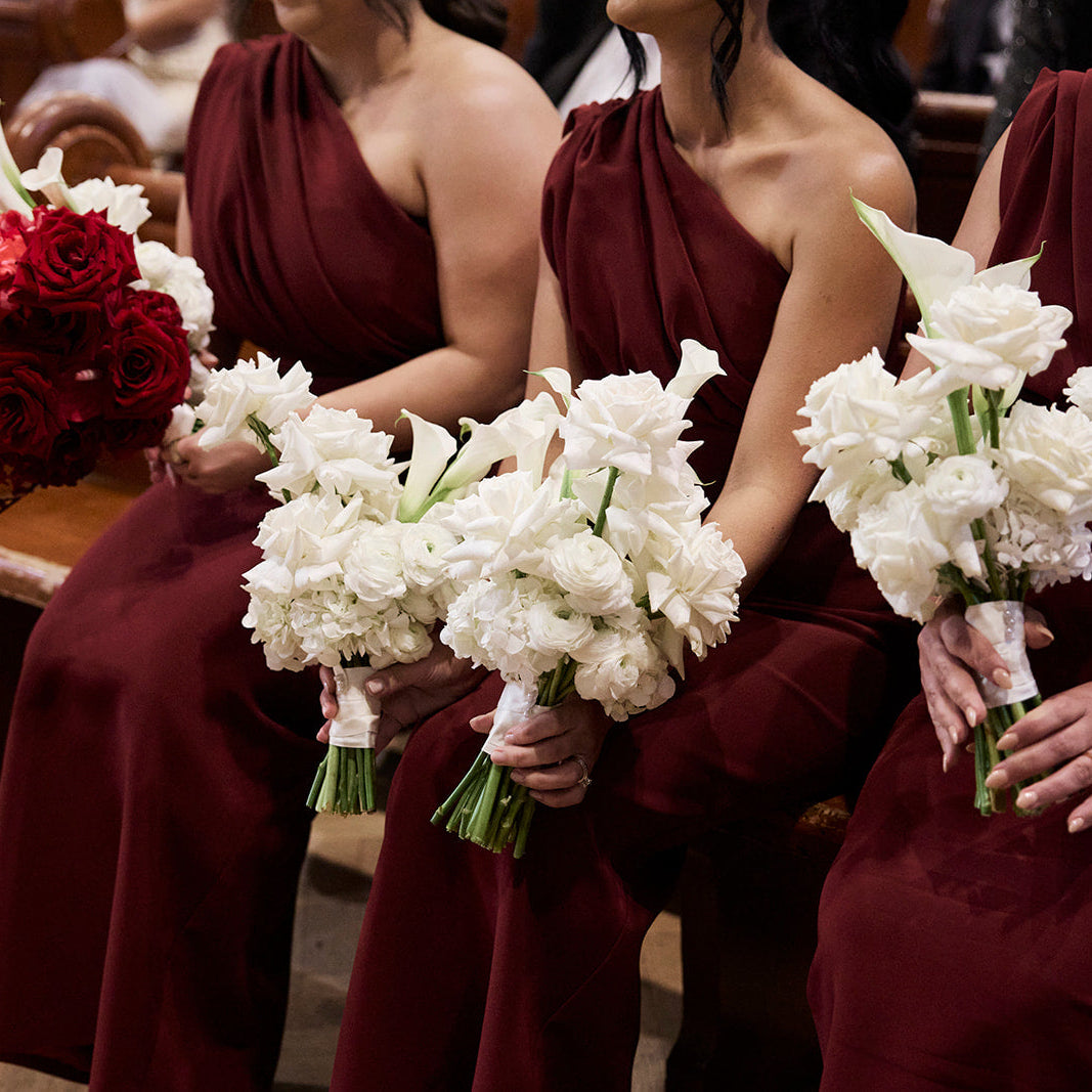 Bridesmaids seated in a church holding white flower bouquets, wearing matching deep burgundy one-shoulder dresses. One bridesmaid on the left holds a contrasting red rose bouquet.