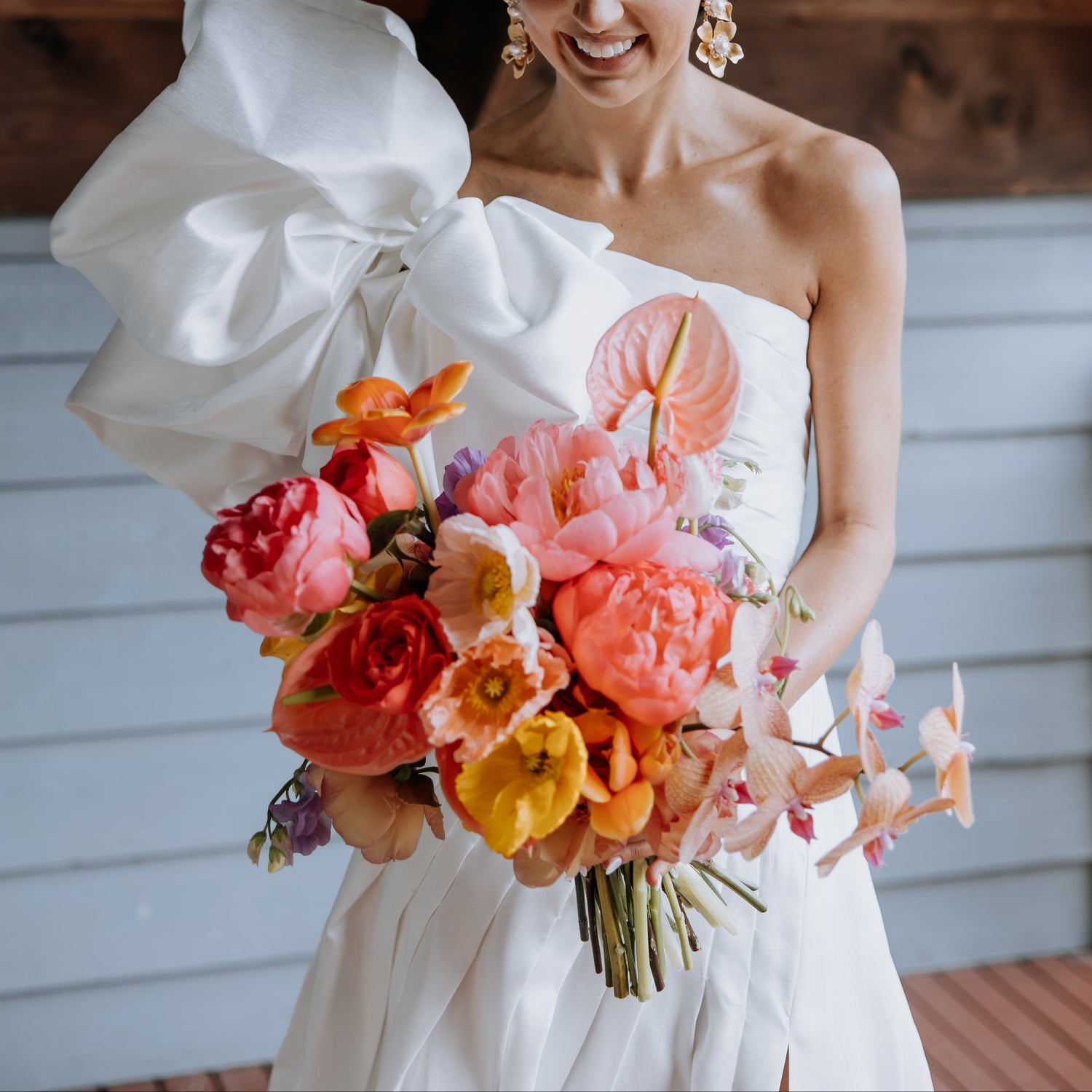 A smiling bride in a white strapless gown with a dramatic oversized bow on the left shoulder holds a vibrant bouquet of pink, orange, and yellow flowers. She wears a white textured headpiece and gold earrings, standing under a wooden ceiling with light blue siding in the background.