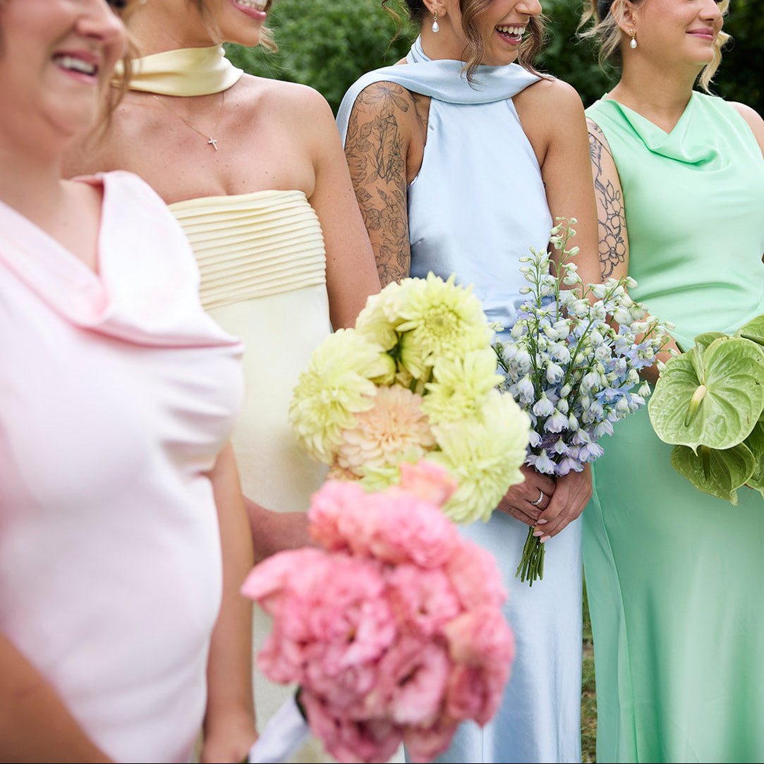 Four women standing outdoors in pastel bridesmaid dresses, holding flower bouquets. The dresses are light pink, pale yellow, light blue, and mint green.