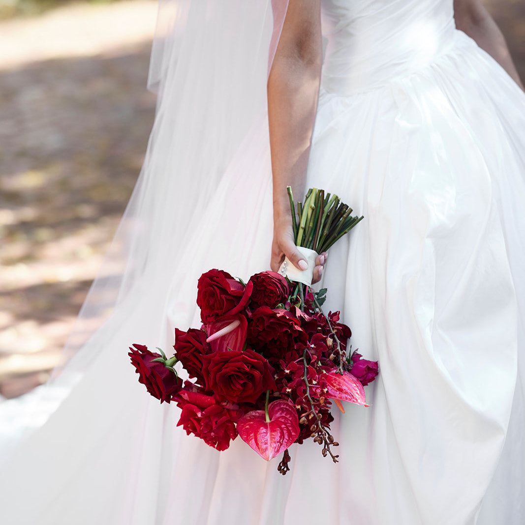 A close-up of a bride in a white wedding gown holding a bouquet of deep red and pink roses outdoors.
