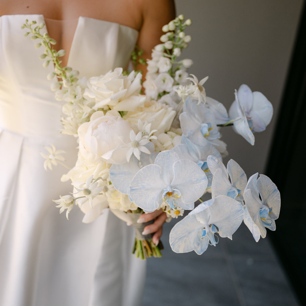 A bride in a strapless white gown holds a large bridal bouquet featuring white and pale blue orchids, white roses, and green foliage.