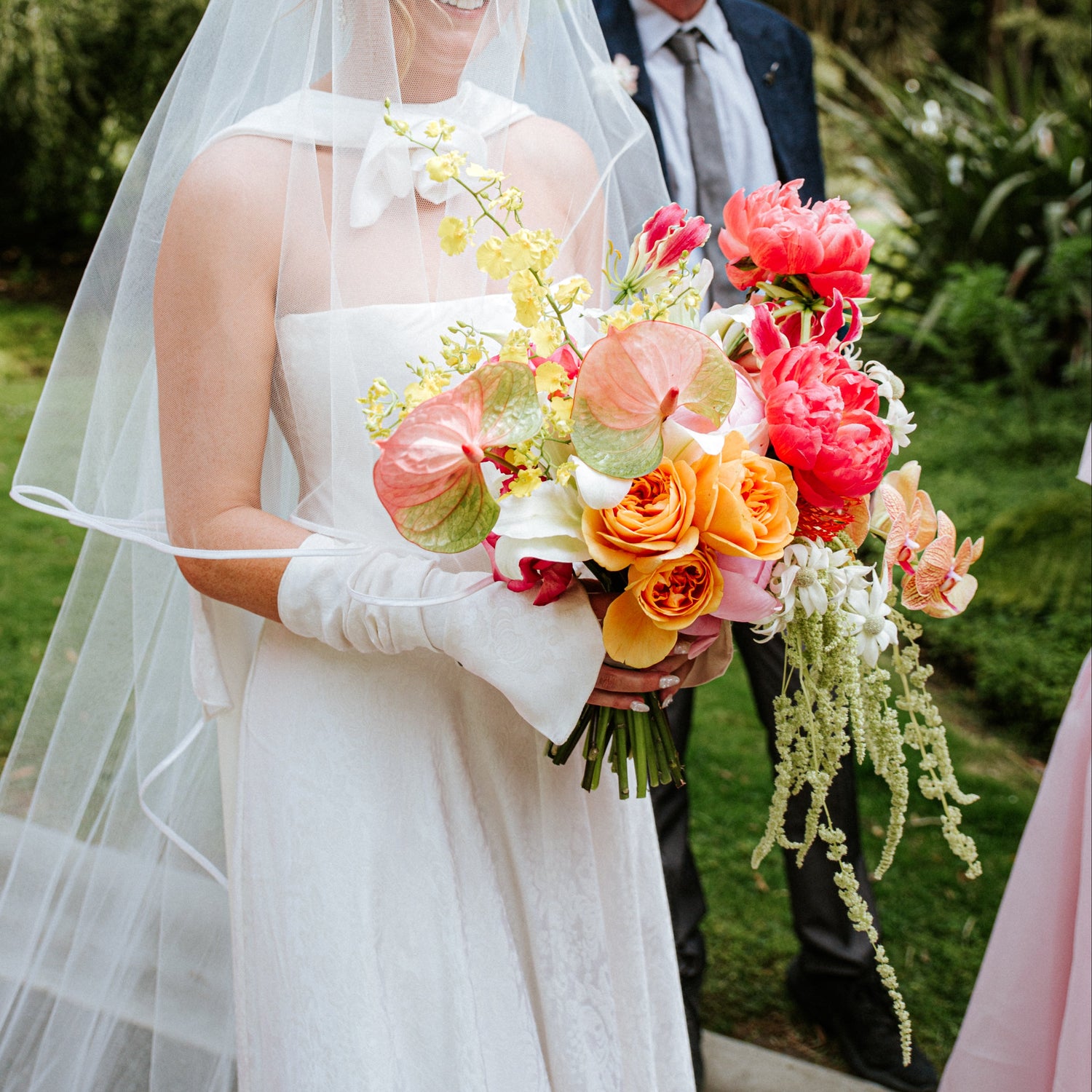 A bride in a white wedding dress and long sheer veil holds a large, vibrant bouquet of pink, orange, and yellow flowers, standing outdoors in a garden with a man in a suit behind her.