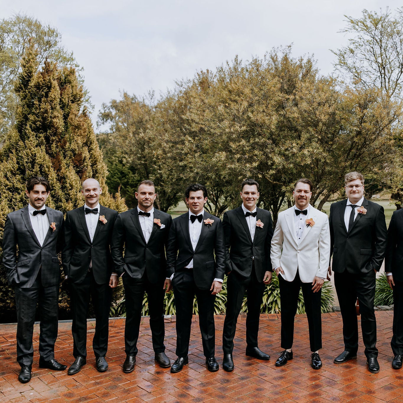 A group of nine men standing outdoors on a brick patio in front of trees. Eight men are wearing black tuxedos with black bow ties and white shirts, while the central man wears a light-colored blazer.