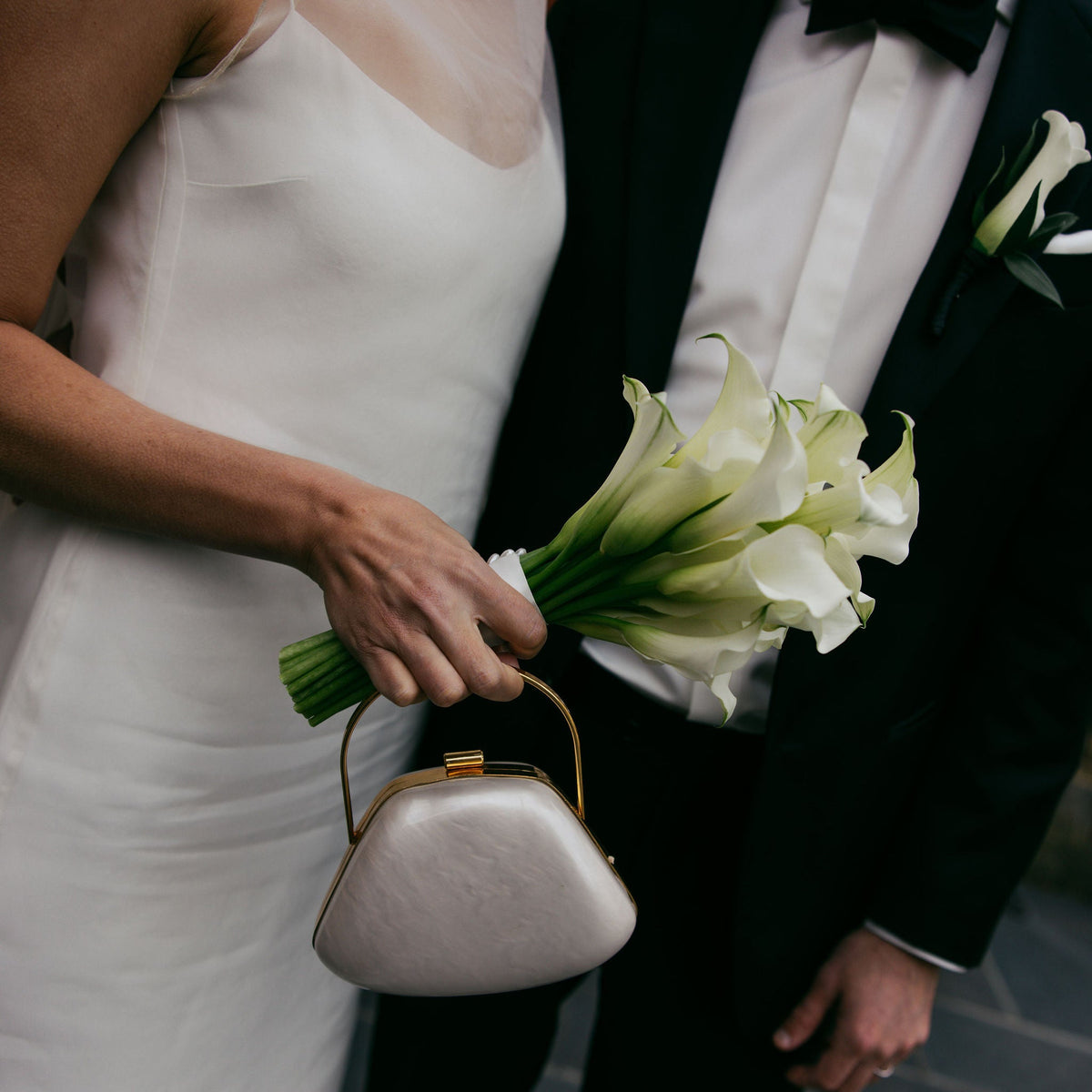 A close-up wedding scene showing a bride in a white sleeveless gown holding a bouquet of white calla lilies and a small metallic clutch, standing beside a groom in a black tuxedo with a bow tie.