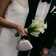 A close-up wedding scene showing a bride in a white sleeveless gown holding a bouquet of white calla lilies and a small metallic clutch, standing beside a groom in a black tuxedo with a bow tie.