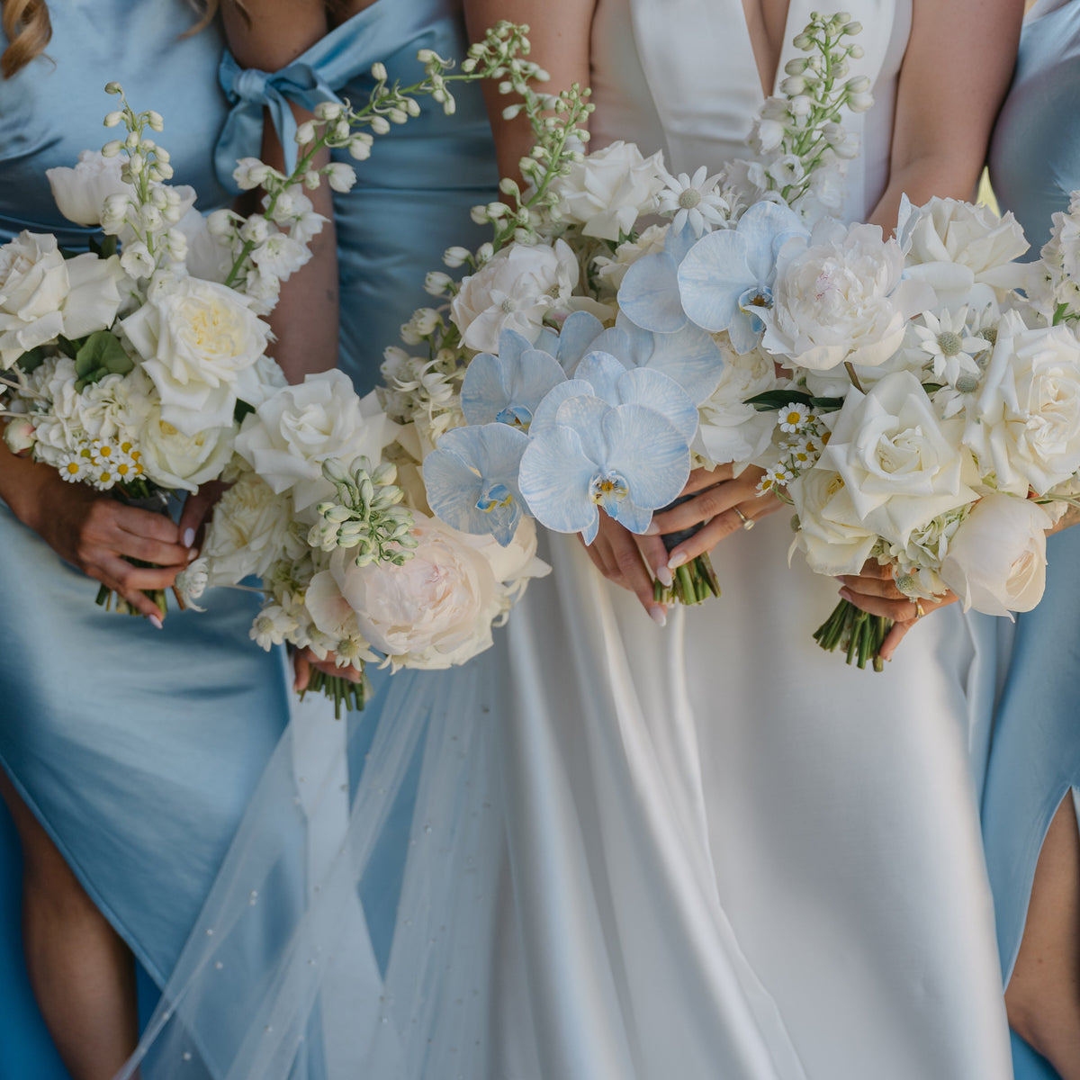 Bridesmaids in light blue satin dresses holding white and pale blue flower bouquets beside a bride in a white satin wedding gown.