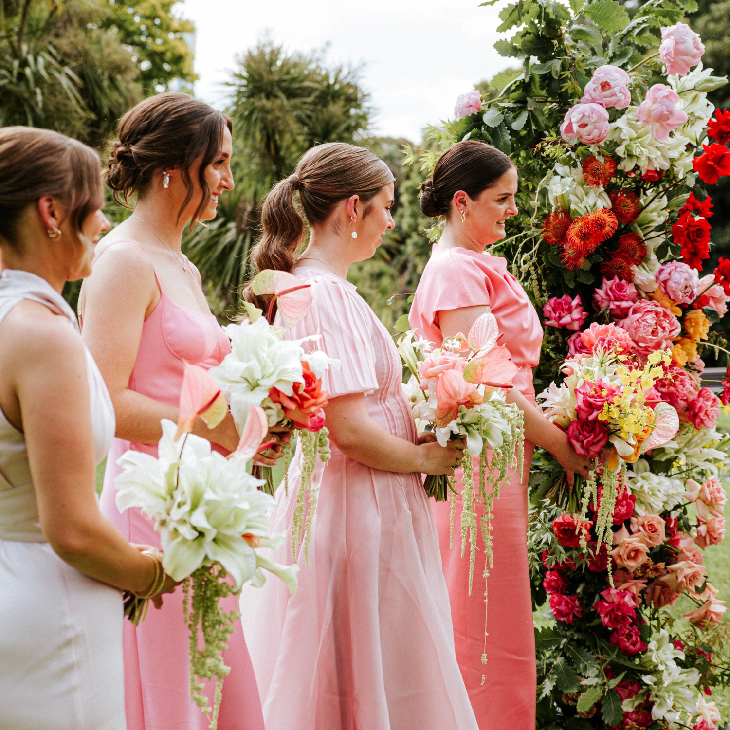 Four bridesmaids in coordinated wedding dresses stand outdoors beside a large floral arch. Two bridesmaids wear white dresses, two wear soft pink dresses, and all hold bouquets.
