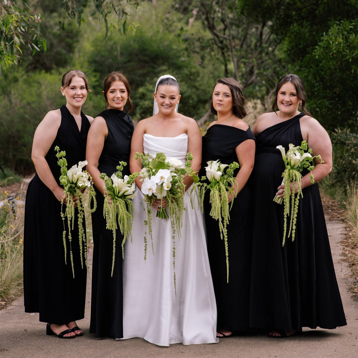 A bride in a white strapless gown stands outdoors with four bridesmaids wearing black dresses, holding white and green floral bouquets on a dirt path with trees in the background.