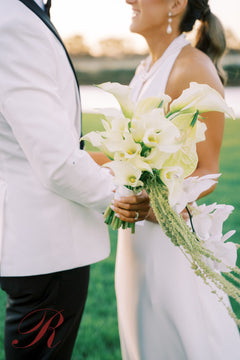 A close-up wedding scene showing a bride in a white sleeveless gown holding a bouquet of white calla lilies and green foliage, standing outdoors with a groom in a white tuxedo jacket and black trousers partially visible.