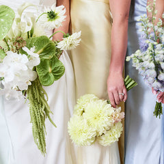 Three bridesmaids in white, cream, and light blue wedding dresses standing together, each holding a floral bouquet. The center bridesmaid wears a cream satin strapless gown and holds a bouquet of pale yellow flowers.