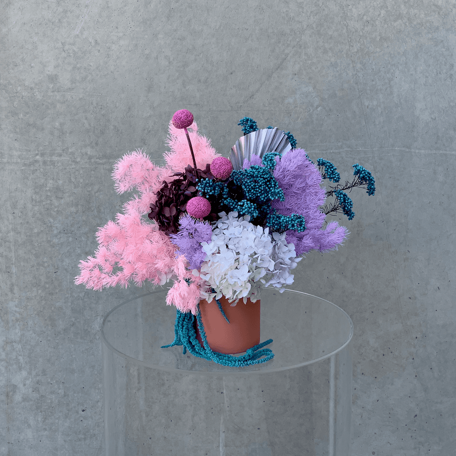Preserved flower arrangement of mixed bright pastel colours displayed in a coral pink vase.